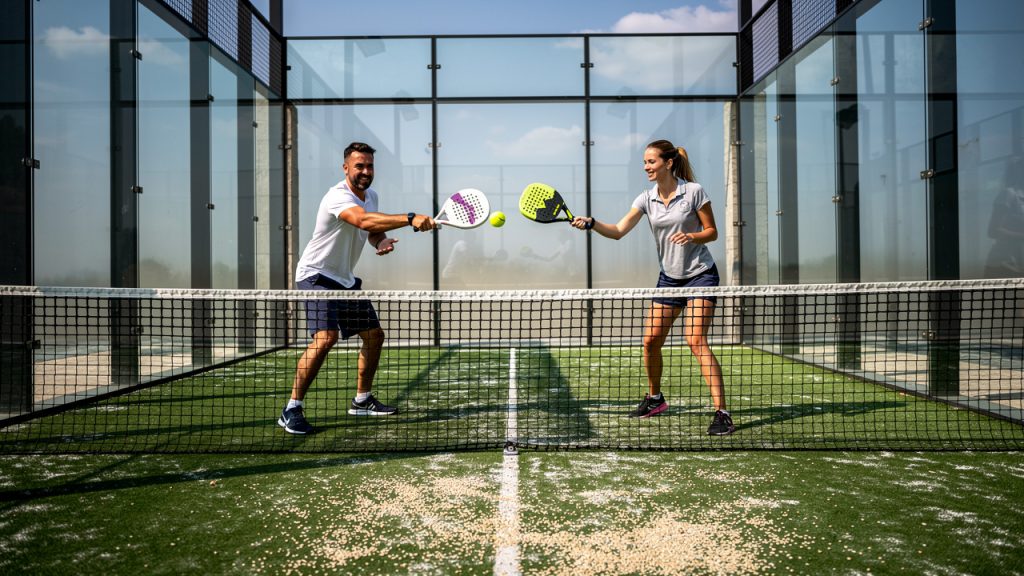 Un homme et une femme débutants jouent au padel sur un court vitré, avec le filet, les parois transparentes et le gazon synthétique sablé visibles en vue large.